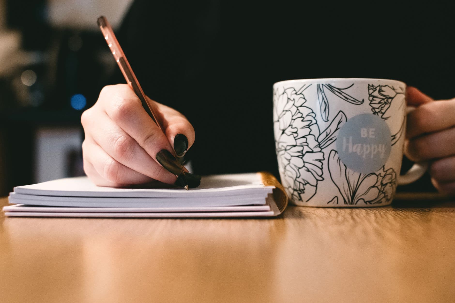 Freelancer redator: como conseguir os primeiros clientes Person writing in a notebook with a floral ceramic mug on a wooden desk.