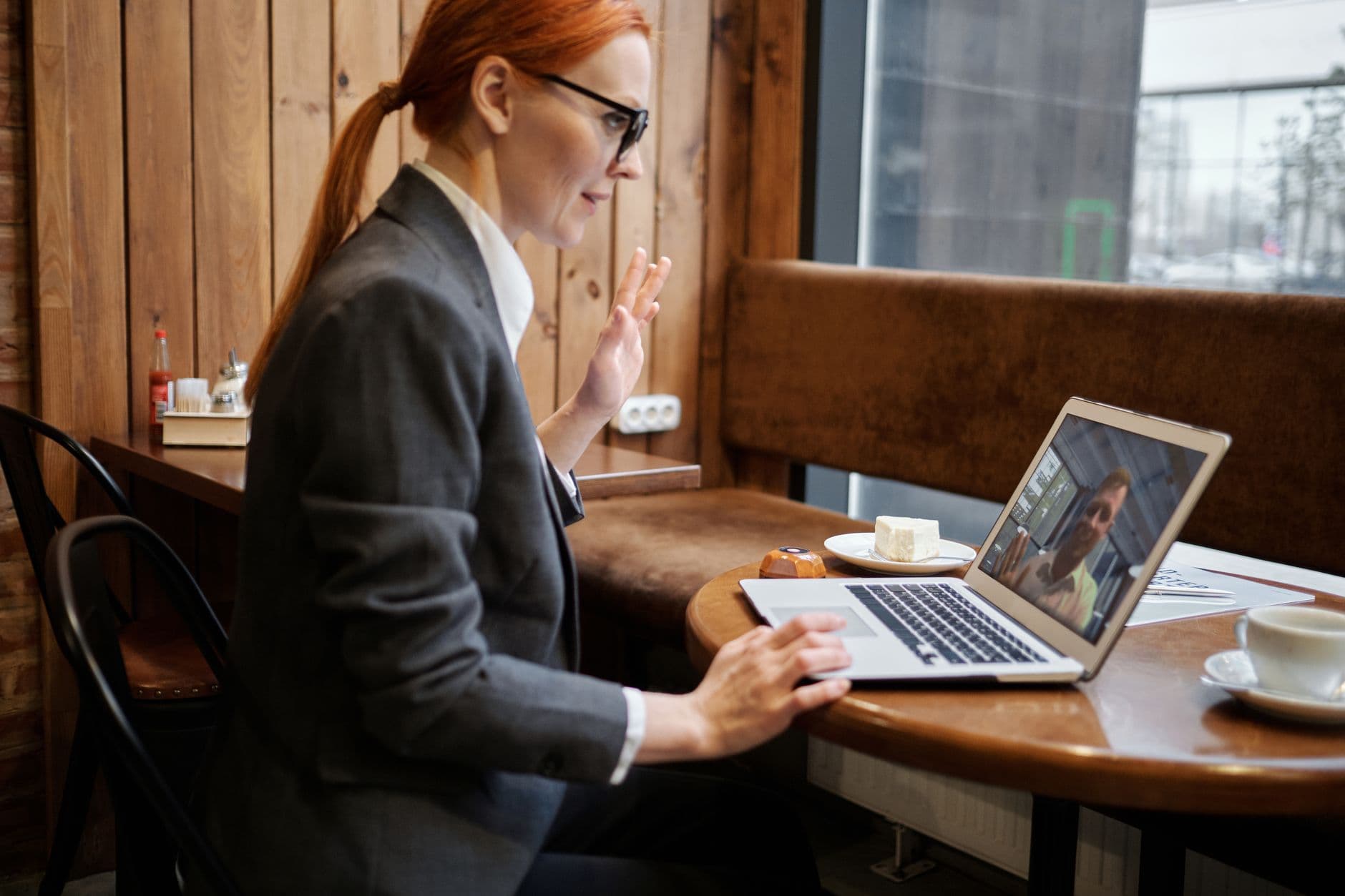 Como fazer networking no trabalho remoto Businesswoman waving during an online meeting in a cozy café setting.