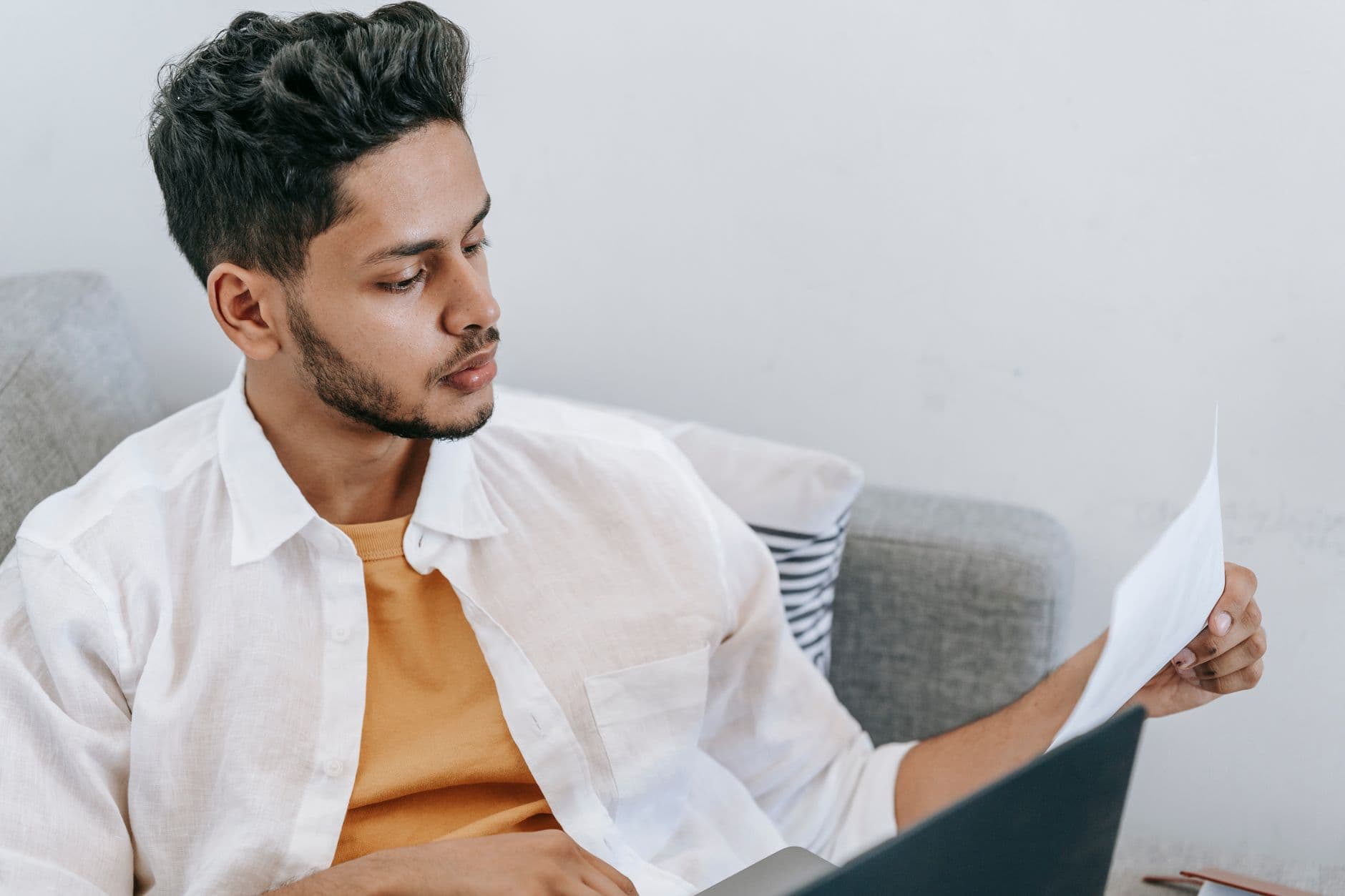 Vagas remotas part-time: onde encontrar Concentrated young ethnic male wearing casual outfit reading document and browsing netbook while resting on cozy couch